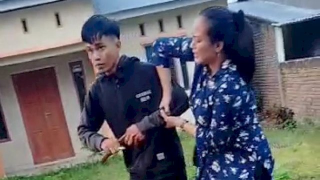 Seorang pemuda dengan balutan sweater hitam dan jeans berwarna hitam memegang senjata tajam jenis Badik. (Tangkapan Layar).